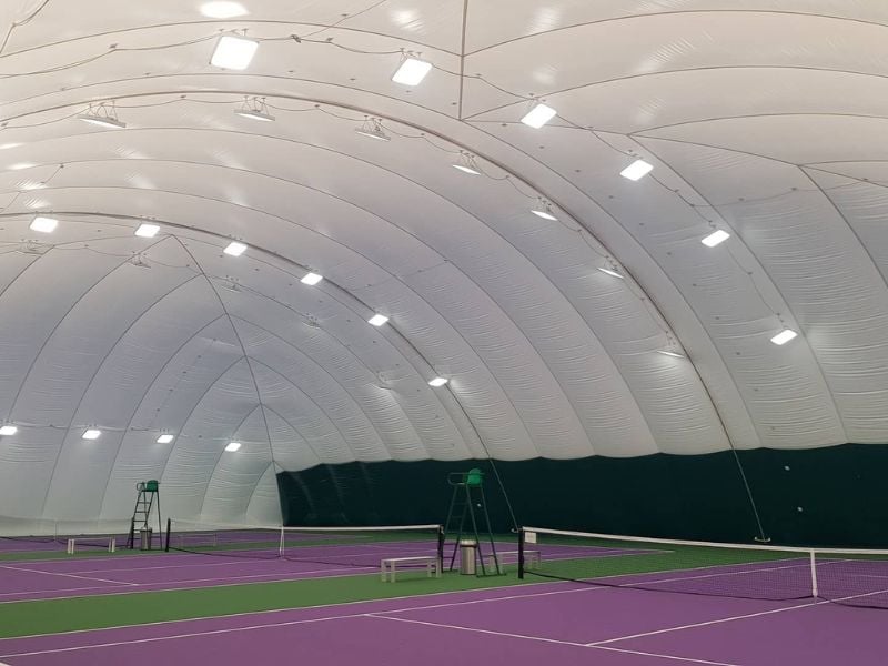 Indoor tennis court in an inflatable dome with a purple surface, lighting fixtures on the ceiling, and green boundary lines.