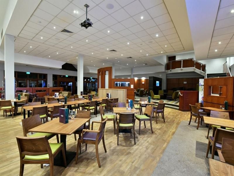 Dining area at DL Northwood Gallery with wooden tables and green-cushioned chairs under ceiling lights, and wooden accents throughout.