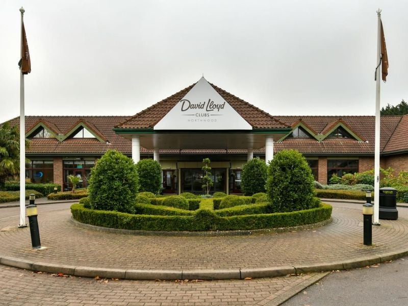 Exterior of David Lloyd Northwood Club with manicured hedges, a triangular roof, and flag poles.