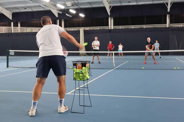 Tennis lesson at David Lloyd Southampton indoor court, featuring an instructor and adult participants practising their swings.