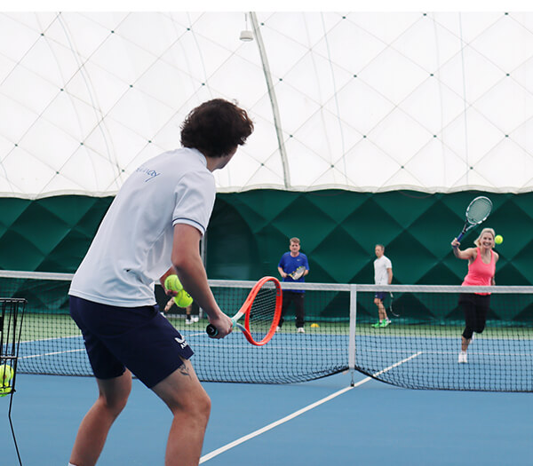 Indoor tennis match at DL Sidcup, showing a player with an orange racket ready to return a serve.