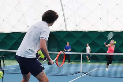 Indoor tennis match at DL Sidcup, showing a player with an orange racket ready to return a serve.