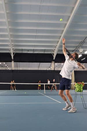 Tennis coach on an indoor court in Southampton, in the middle of serving the ball, with players practising in the background.