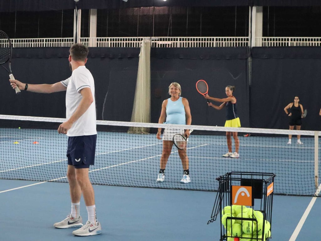 Tennis coaching session on an indoor court with a coach guiding players, including a woman in blue and a woman in a yellow skirt, with a rack of tennis balls.