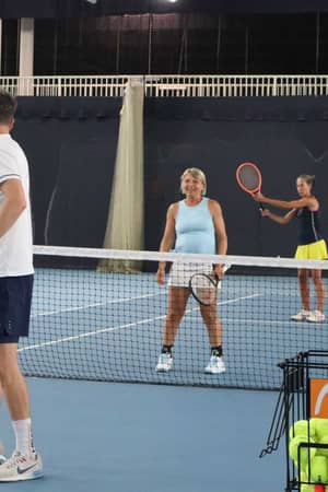 Tennis coaching session on an indoor court with a coach guiding players, including a woman in blue and a woman in a yellow skirt, with a rack of tennis balls.