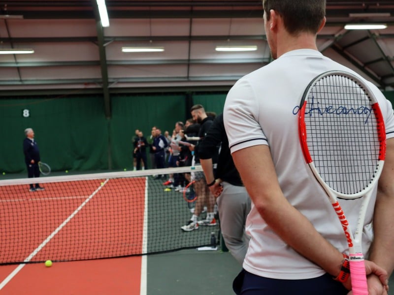 Tennis coach in a white shirt holding a racket stands by the net on a red indoor court, observing a group coaching session.