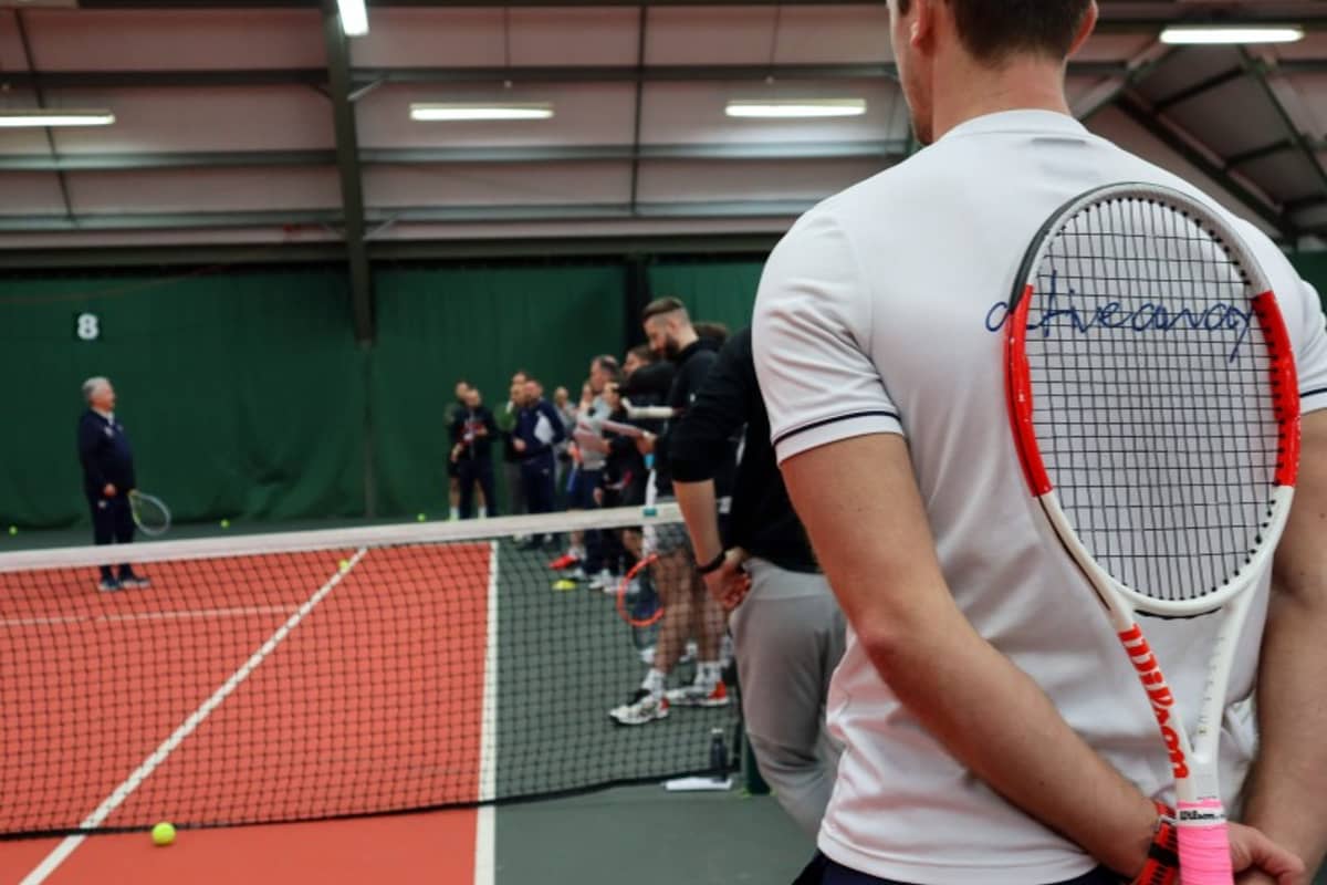 Tennis coach in a white shirt holding a racket stands by the net on a red indoor court, observing a group coaching session.