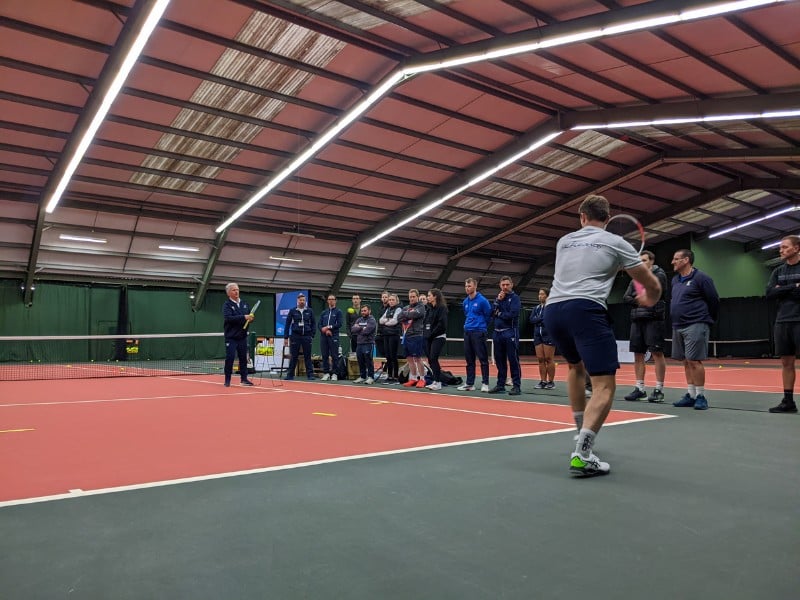 Indoor tennis training at David Lloyd West Bridgford with players watching a coach demonstrate a drill.