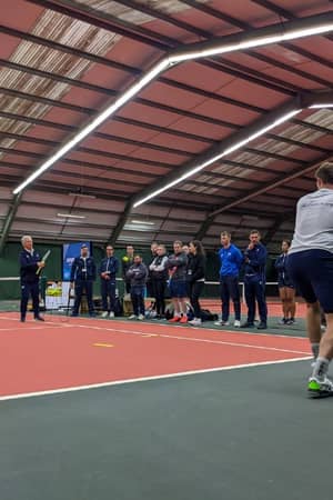 Indoor tennis training at David Lloyd West Bridgford with players watching a coach demonstrate a drill.