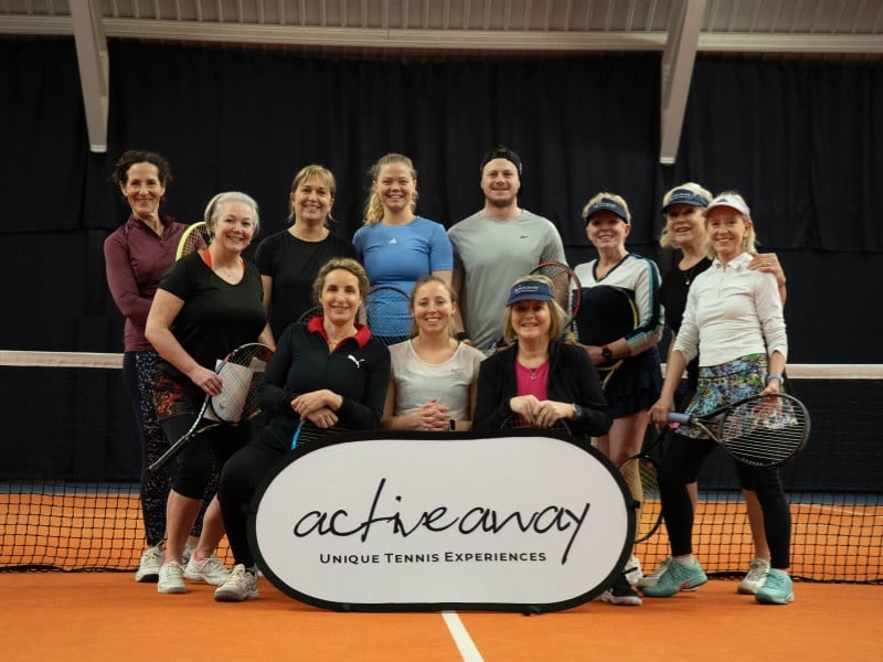 Group of tennis players on indoor court with Active Away sign, featuring nine women and one man holding tennis racquets.