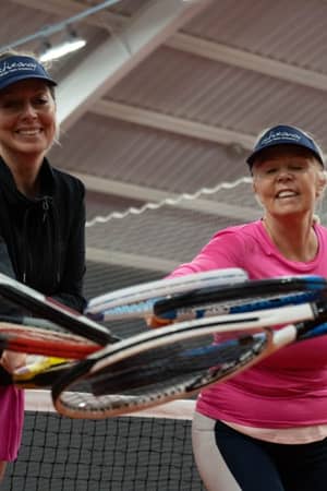 Three women in tennis outfits with visors, joining their racquets together inside an indoor tennis court, expressing teamwork and camaraderie.