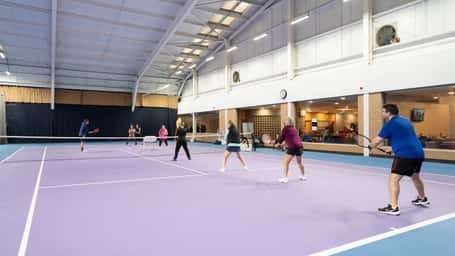 Indoor tennis facility with players engaged in a doubles match on a purple court.