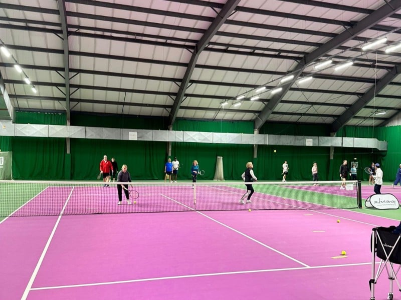 Indoor tennis court in Woking with players engaged in a doubles match on a pink surface and green walls.