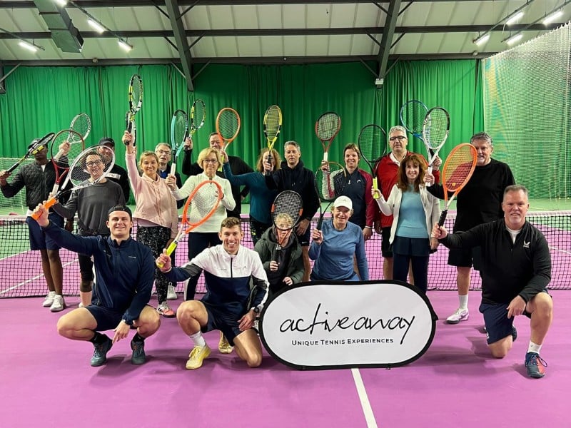 Group of tennis players at DL Woking holding rackets and a banner for Active Away Unique Tennis Experiences on an indoor court.