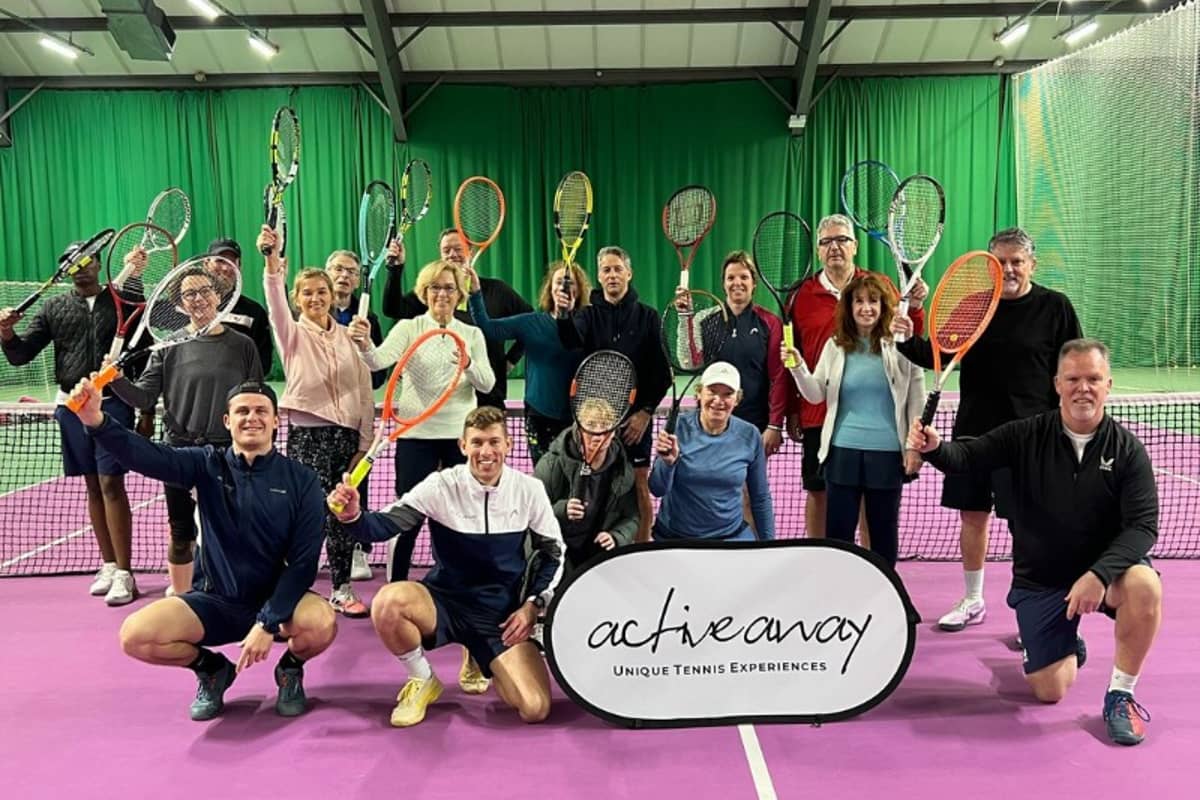 Group of tennis players at DL Woking holding rackets and a banner for Active Away Unique Tennis Experiences on an indoor court.