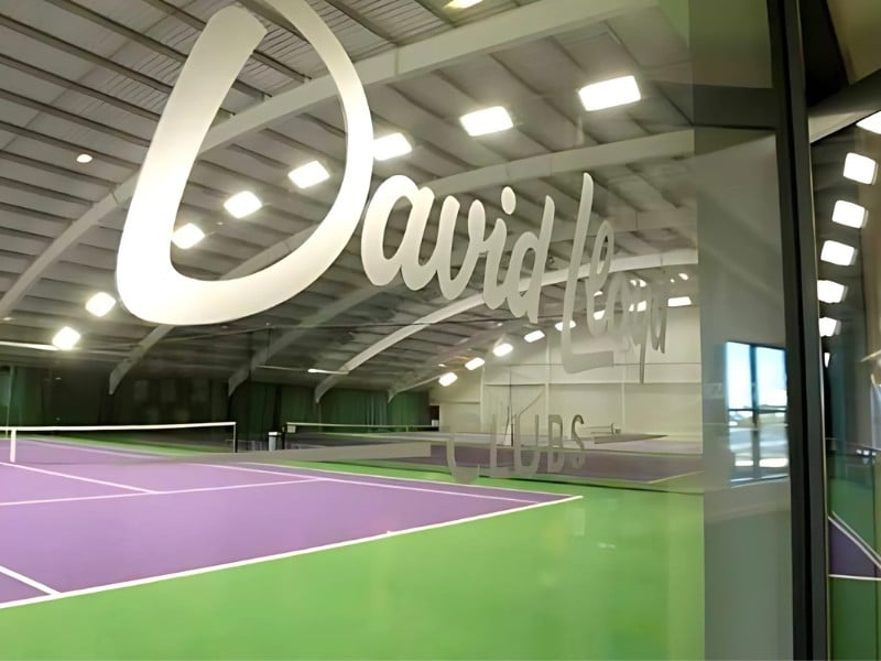 Indoor tennis court at David Lloyd Clubs, Woking, with purple and green floor and David Lloyd Clubs logo on glass window.