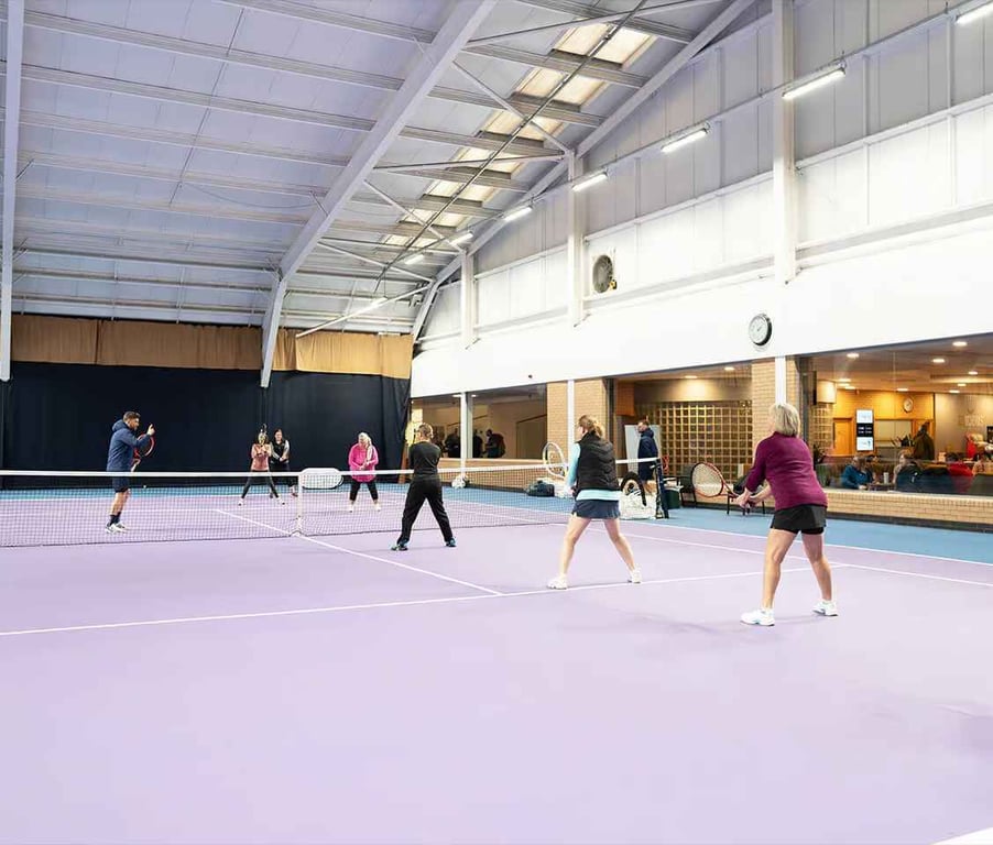 Clients participating in indoor tennis drills at DL Finchley on a spacious, well-lit court with seating areas.