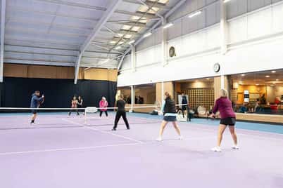 Clients participating in indoor tennis drills at DL Finchley on a spacious, well-lit court with seating areas.