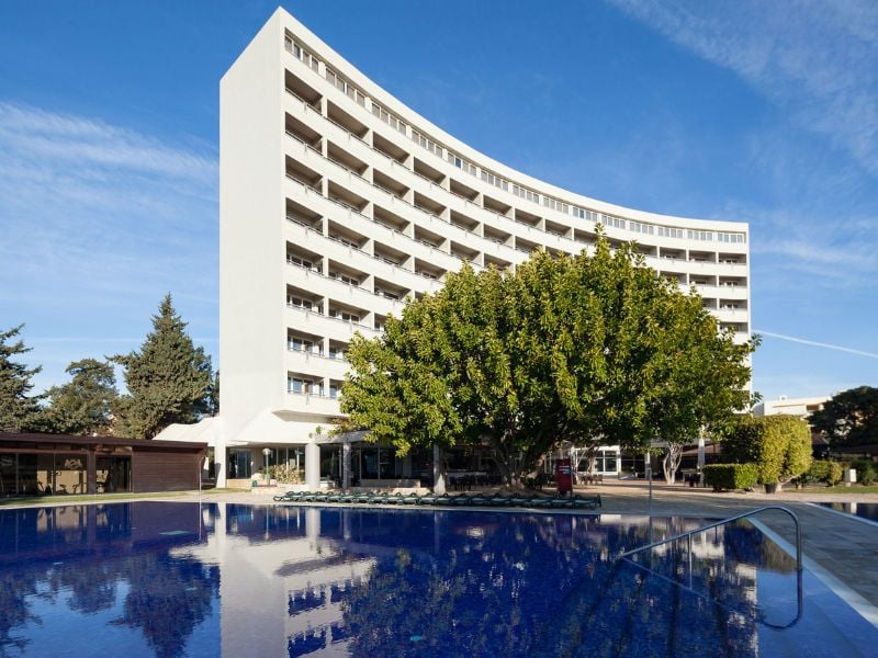 Exterior view of Dom Pedro Hotel with a modern curved design, multiple balconies, and a pool in front, surrounded by trees.