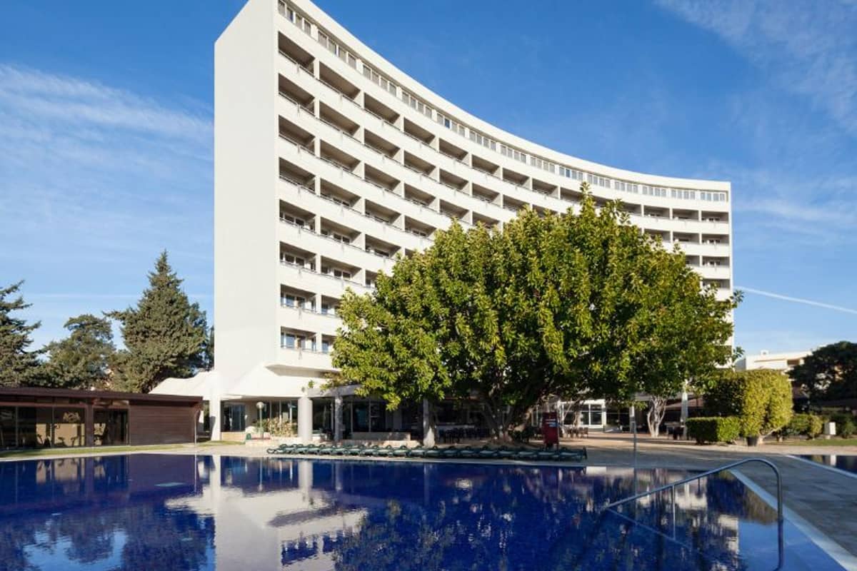 Exterior view of Dom Pedro Hotel with a modern curved design, multiple balconies, and a pool in front, surrounded by trees.