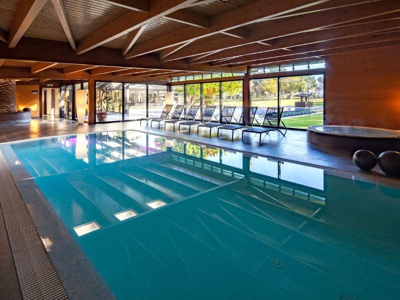 Indoor pool at Dom Pedro Gallery with wooden beams, large windows, and sun loungers, offering a view of greenery outside.