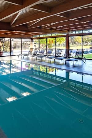 Indoor pool at Dom Pedro Gallery with wooden beams, large windows, and sun loungers, offering a view of greenery outside.