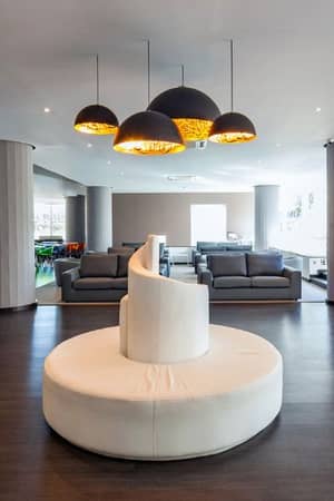 Modern lounge area in Dom Pedro Gallery with a central circular white sofa, grey armchairs, pendant lights, and potted plants.