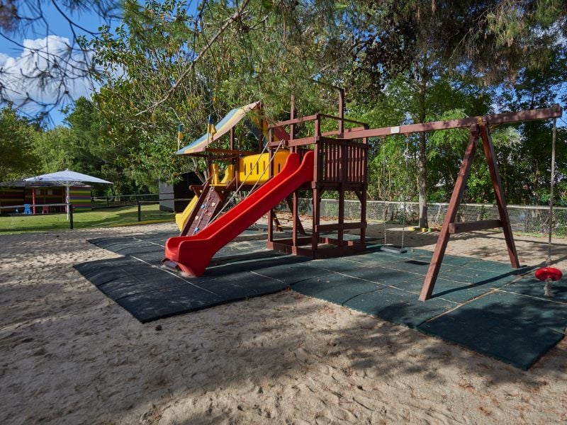 Dom Pedro Gallery playground with a colourful play structure, red slide, and swings, set amidst green trees.