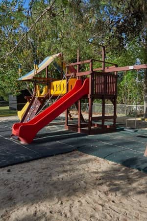 Dom Pedro Gallery playground with a colourful play structure, red slide, and swings, set amidst green trees.