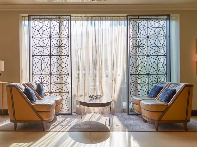 Lounge area at Dona Filipa Hotel with beige armchairs, blue cushions, a glass coffee table, and geometric-patterned screens.