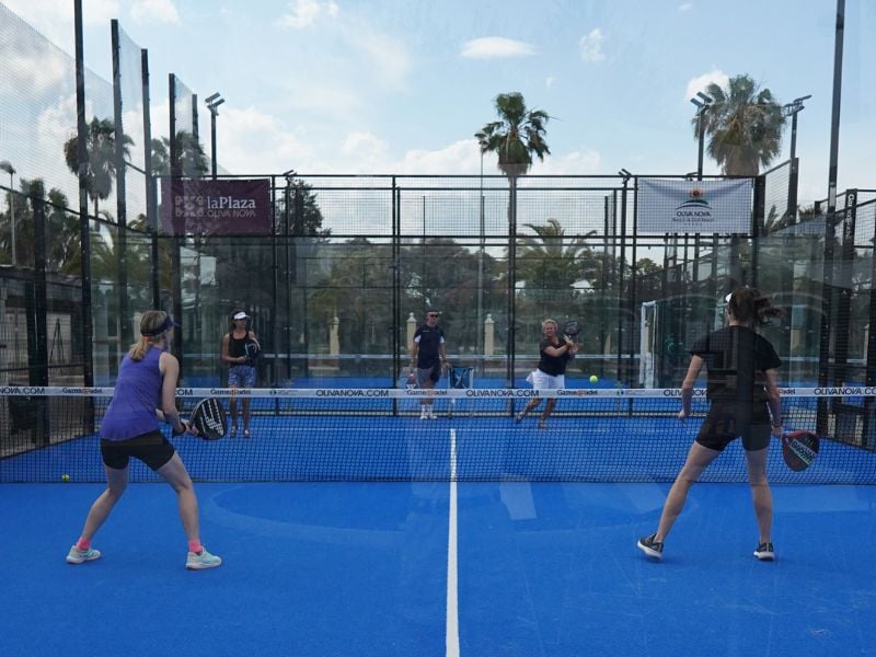 Players in action on a blue padel court at Dona Filipa, surrounded by glass walls and palm trees, with spectators watching.