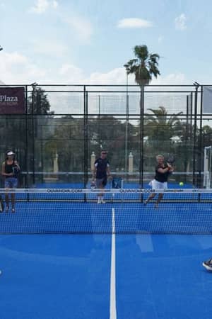 Players in action on a blue padel court at Dona Filipa, surrounded by glass walls and palm trees, with spectators watching.