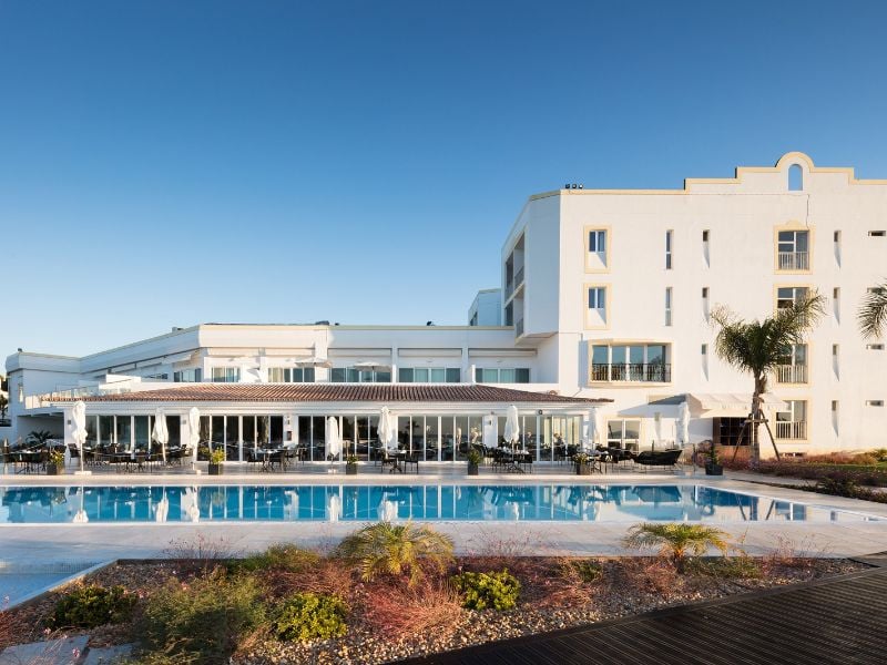 Dona Filipa Hotel pool area, showcasing a large pool with surrounding modern white architecture, palm trees, and a clear blue sky.