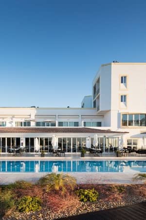 Dona Filipa Hotel pool area, showcasing a large pool with surrounding modern white architecture, palm trees, and a clear blue sky.