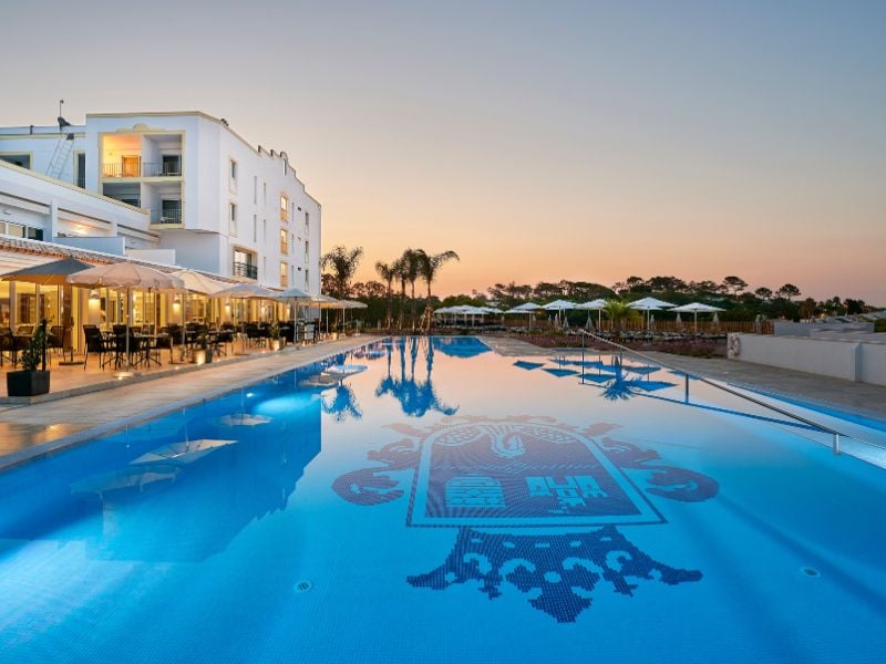 Tranquil outdoor swimming pool at Dona Filipa Padel Gallery reflecting the sunset, with hotel and palm trees in the background.