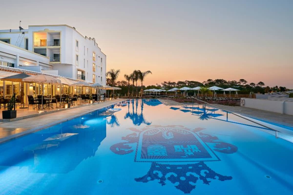Tranquil outdoor swimming pool at Dona Filipa Padel Gallery reflecting the sunset, with hotel and palm trees in the background.