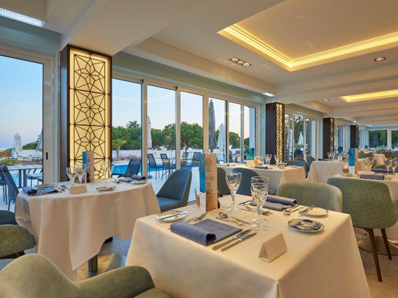 Interior of the Dona Filipa Hotel restaurant with tables set for fine dining, overlooking the pool and lush greenery through expansive windows.