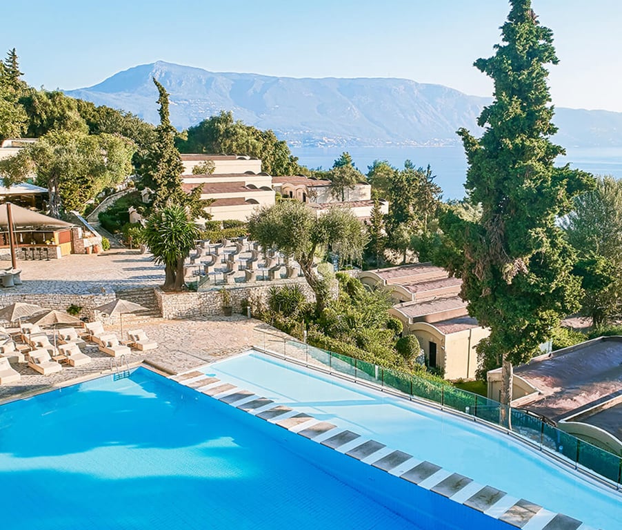 View of Daphnila Bay resort, showcasing a blue pool, sun loungers, and villas with a mountain and sea in the background.