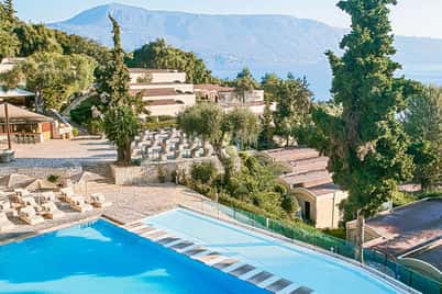 View of Daphnila Bay resort, showcasing a blue pool, sun loungers, and villas with a mountain and sea in the background.