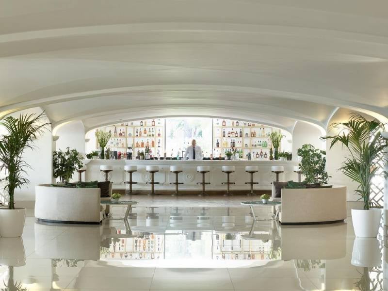 Daphnila Bay Bar with a modern white design, curved ceilings, well-stocked shelves, bar stools, and potted plants.