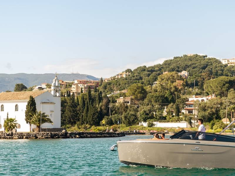 View of a cruise in Daphnila Bay with a backdrop of green hills, buildings, and a white chapel by the water.