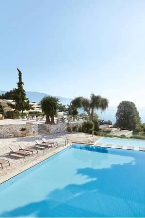 Swimming pool at Daphnila Bay surrounded by lounge chairs and greenery, overlooking the sea and a clear sky.