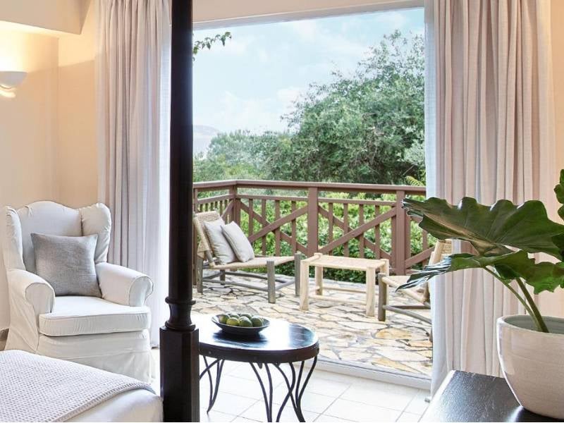 Balcony view from a room at Daphnila Bay, with a white armchair, round table, and lush greenery outside.