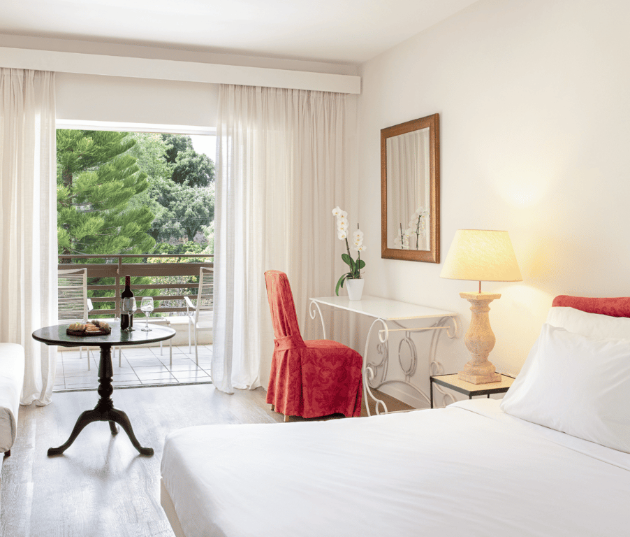 Interior of Daphnila Bay Superior Room with a white double bed, red chair, round table, and balcony overlooking garden.