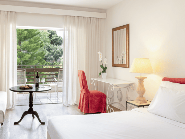 Interior of Daphnila Bay Superior Room with a white double bed, red chair, round table, and balcony overlooking garden.