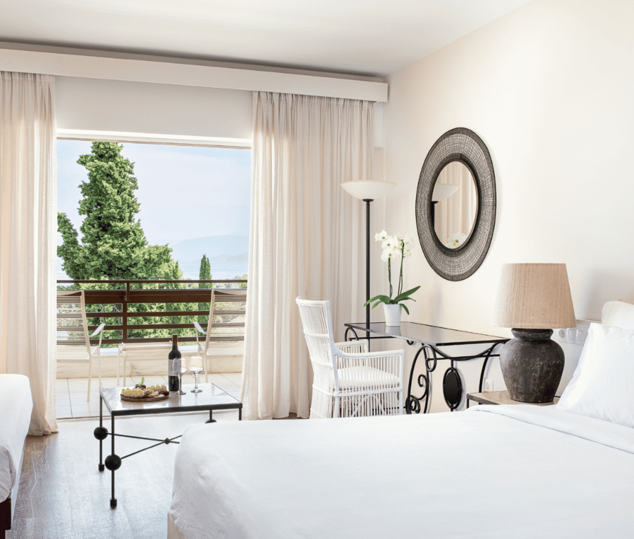 Superior room at Daphnila Bay with a sea view and private balcony, featuring white linens, modern decor, a table with wine, and large windows showcasing the sea and greenery.