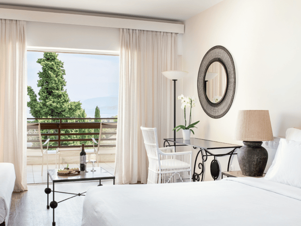 Superior room at Daphnila Bay with a sea view and private balcony, featuring white linens, modern decor, a table with wine, and large windows showcasing the sea and greenery.