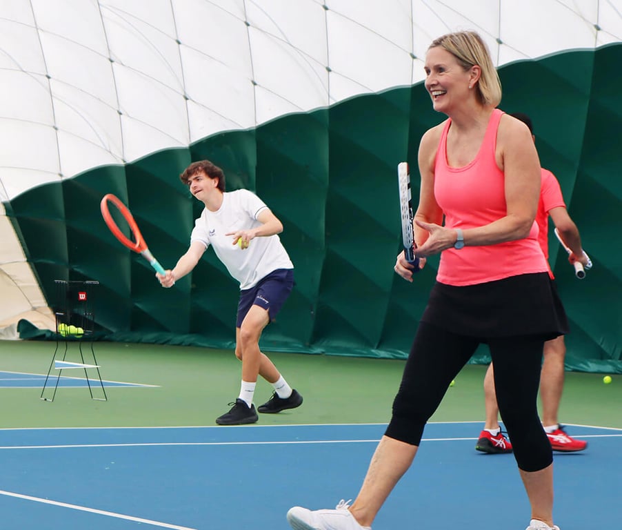 Woman in orange tank top playing tennis indoors at David Lloyd Bristol with a young male player in the background.