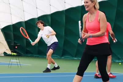 Woman in orange tank top playing tennis indoors at David Lloyd Bristol with a young male player in the background.