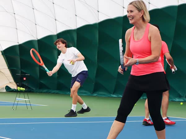 Woman in orange tank top playing tennis indoors at David Lloyd Bristol with a young male player in the background.
